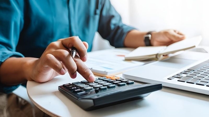 Person berechnet Finanzen mit Taschenrechner und Laptop am Schreibtisch