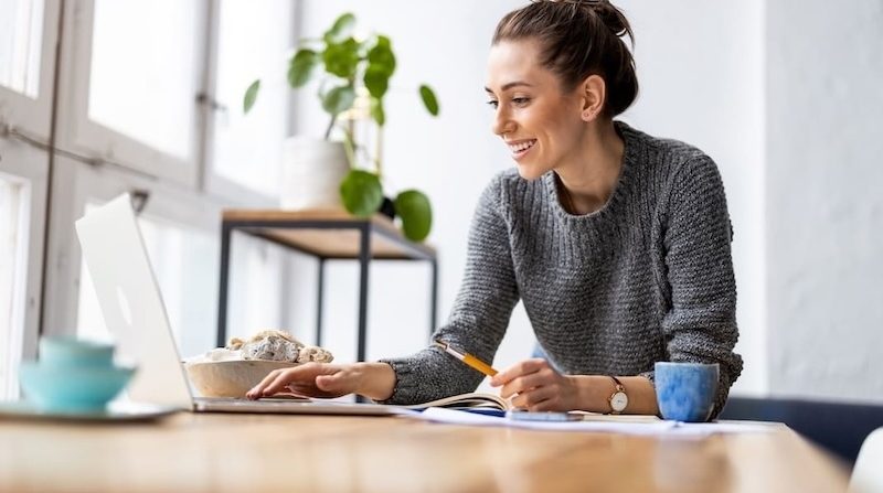 Junge Frau arbeitet lächelnd am Laptop an einem hellen Schreibtisch im Homeoffice
