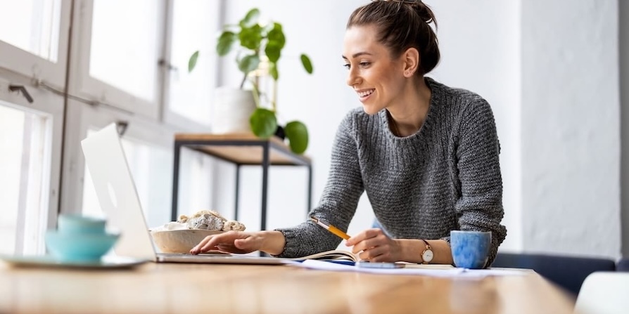 Junge Frau arbeitet lächelnd am Laptop an einem hellen Schreibtisch im Homeoffice