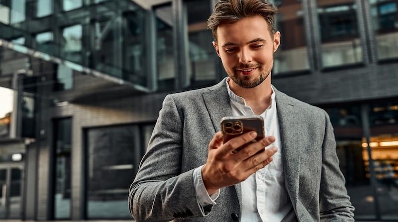 Junger Geschäftsmann schaut lächelnd auf sein Smartphone vor modernem Bürogebäude