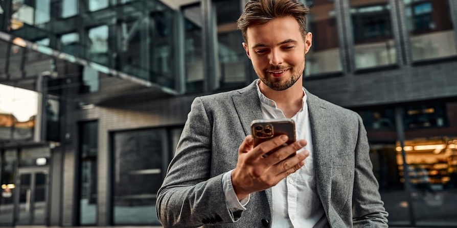 Junger Geschäftsmann schaut lächelnd auf sein Smartphone vor modernem Bürogebäude