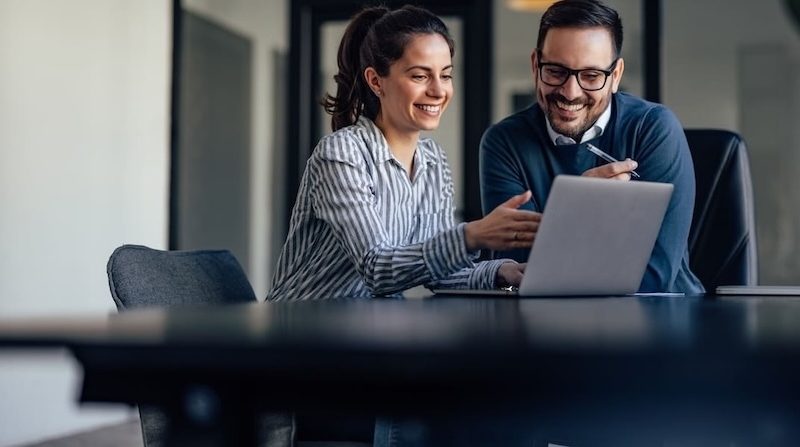 Zwei Kollegen arbeiten lächelnd gemeinsam an einem Laptop im modernen Büro