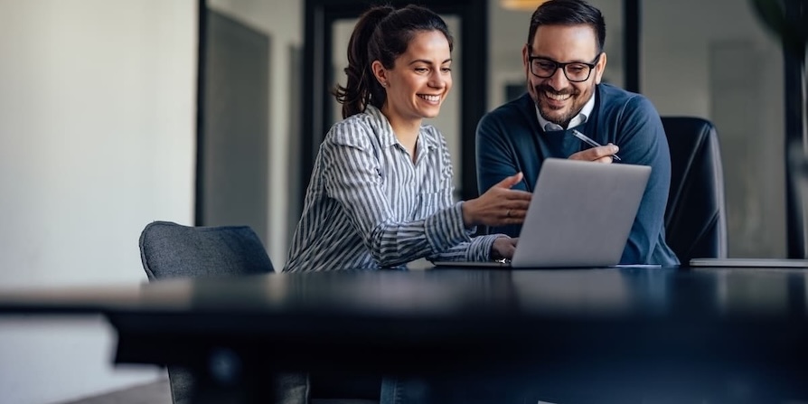 Zwei Kollegen arbeiten lächelnd gemeinsam an einem Laptop im modernen Büro