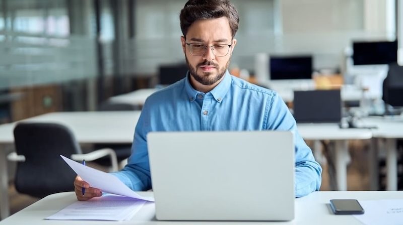 Mann arbeitet konzentriert am Laptop und prüft Unterlagen im modernen Büro