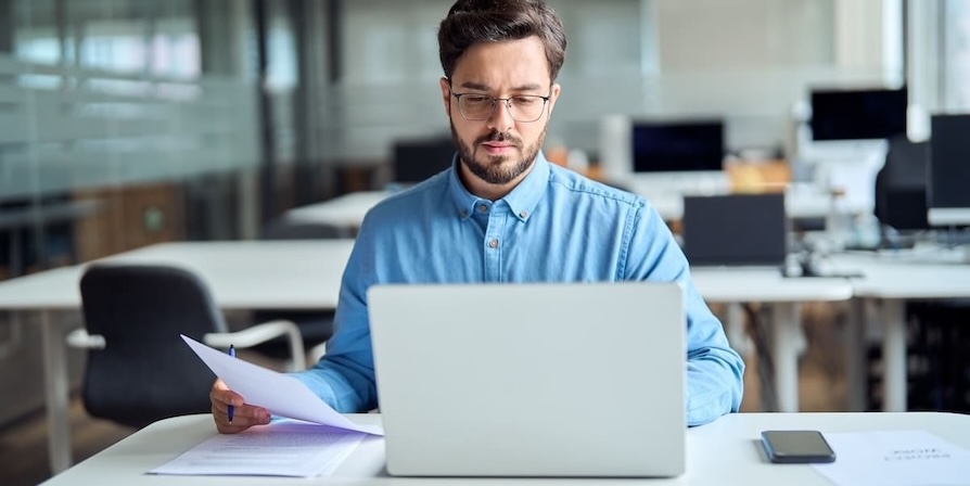 Mann arbeitet konzentriert am Laptop und prüft Unterlagen im modernen Büro