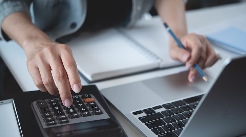 Hände einer Person beim Berechnen und Dokumentieren von Finanzdaten mit Taschenrechner, Notizbuch und Laptop – symbolisiert präzise Analyse- und Büroarbeit.