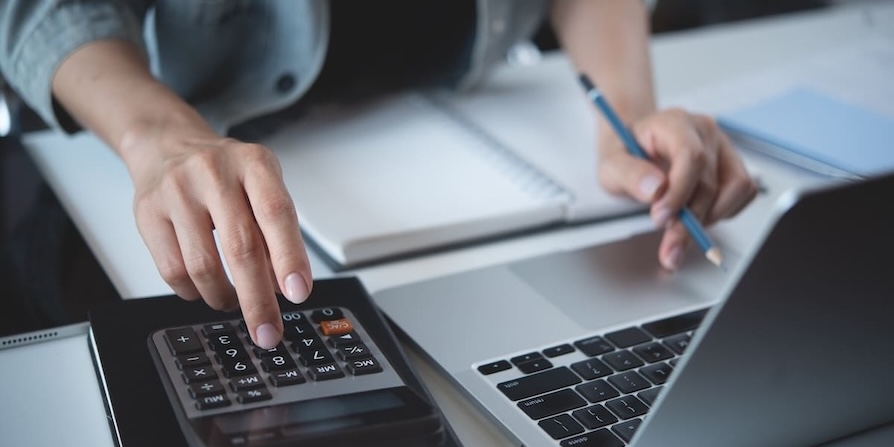 Hände einer Person beim Berechnen und Dokumentieren von Finanzdaten mit Taschenrechner, Notizbuch und Laptop – symbolisiert präzise Analyse- und Büroarbeit.