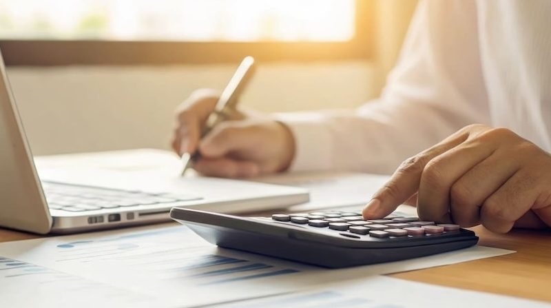 Nahaufnahme einer Hand, die auf einem Taschenrechner tippt, während im Hintergrund Notizen und ein Laptop zu sehen sind – symbolisiert professionelle Finanz- und Analysearbeit.