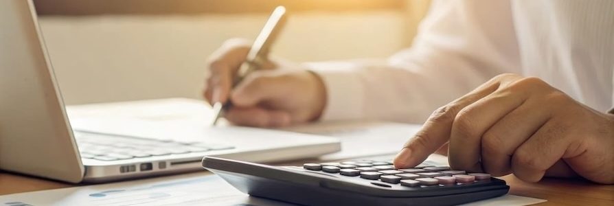 Nahaufnahme einer Hand, die auf einem Taschenrechner tippt, während im Hintergrund Notizen und ein Laptop zu sehen sind – symbolisiert professionelle Finanz- und Analysearbeit.