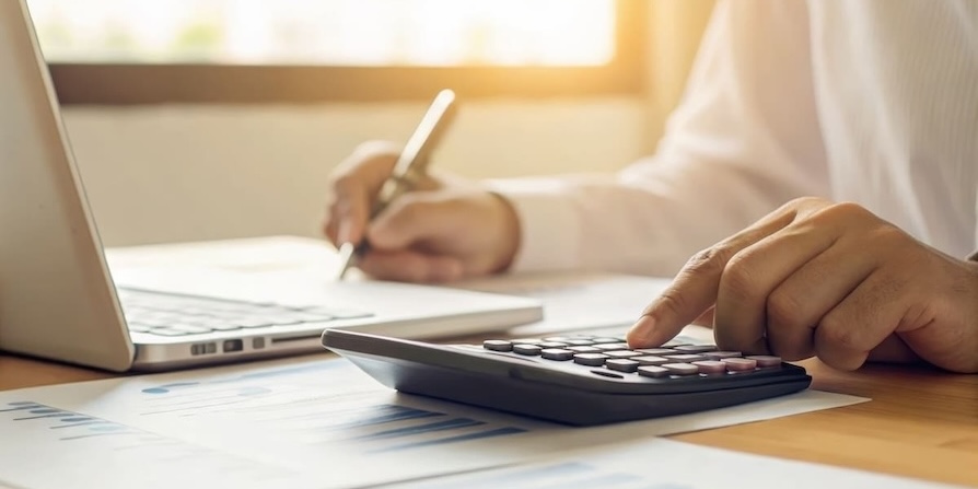 Nahaufnahme einer Hand, die auf einem Taschenrechner tippt, während im Hintergrund Notizen und ein Laptop zu sehen sind – symbolisiert professionelle Finanz- und Analysearbeit.