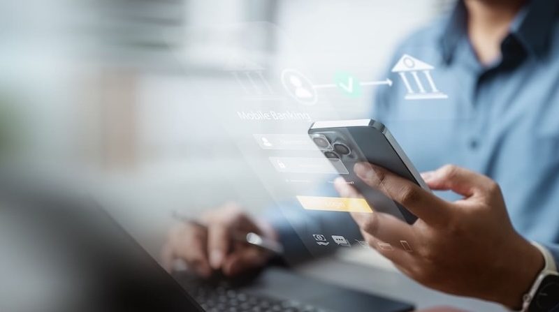 Nahaufnahme einer Person, die ein Smartphone hält und am Laptop arbeitet. Überblendete Grafiken visualisieren Mobile Banking, Login-Daten und digitale Finanzprozesse.