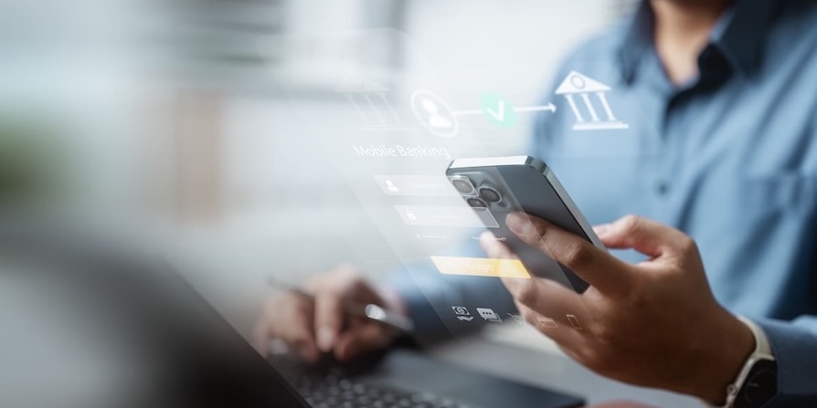 Nahaufnahme einer Person, die ein Smartphone hält und am Laptop arbeitet. Überblendete Grafiken visualisieren Mobile Banking, Login-Daten und digitale Finanzprozesse.